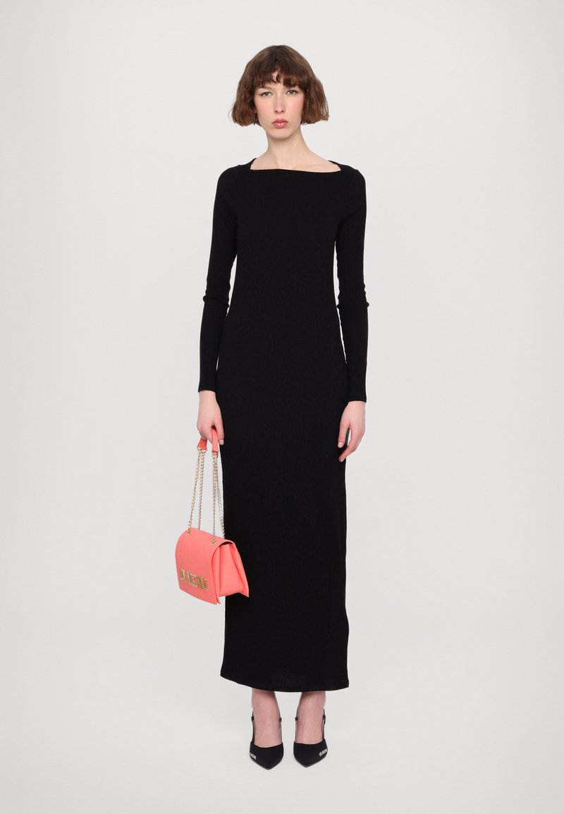 Abito lungo nero con maniche a lunghe e scollatura quadrata, dalla silhouette aderente. La modella tiene una borsa corallo con tracolla a catena e indossa tacchi neri.