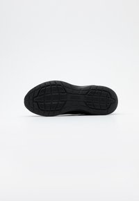 Semelle de chaussure de sport noire en matière caoutchoutée texturée avec un motif de bande de roulement pour l’adhérence et un petit détail de logo en relief.