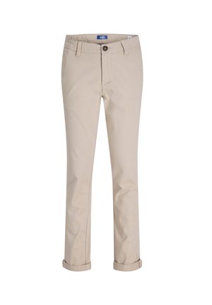 Garçon souriant aux cheveux châtain clair portant une chemise noire boutonnée, un pantalon beige et des baskets blanches avec des rayures noires, debout sur un fond blanc.