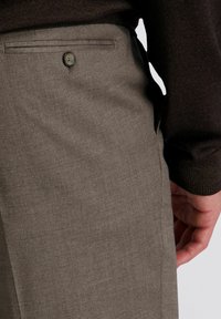 Close-up van de hand van een persoon nabij de achterzak van een grijzige pantalon, die een donker langslavend top draagt.