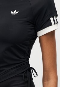 Μαύρο αθλητικό τοπ με ραφές που μαζεύουν στις πλαϊνές λεπτομέρειες, λευκά μανσέτες και μικρό λευκό λογότυπο. Κατασκευασμένο από λείο, ελαφρύ ύφασμα. Τριπλές ρίγες στα μανίκια.