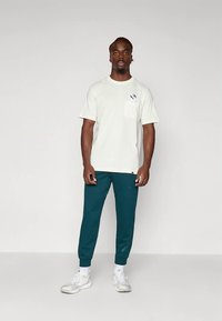 Ανοιχτό μπεζ κοντομάνικο T-shirt με τσέπη στο στήθος, σε συνδυασμό με πετρόλ παντελόνι jogger και λευκά αθλητικά παπούτσια. Απλός σχεδιασμός, άνετη εφαρμογή.