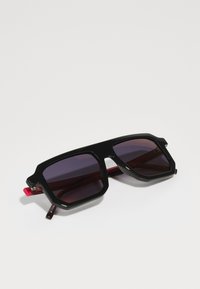 Ochelari de soare dreptunghiulari negri cu lentile întunecate, având un cadru îndrăzneț și unghiular și accente roșii pe brațe. Finisaj lucios, design robust.