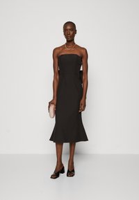 Schwarzes schulterfreies Kleid mit eng anliegendem Oberteil, ausgestelltem Saum und strukturiertem Stoff. Kombiniert mit einer rosa Perlenclutch und schwarzen Absatzsandalen.