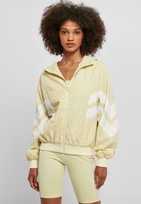 Ljusgul zip-up jacka med huva, prydd med vita chevronränder på ärmarna. Designad med en gatherad nederkant och mjuk textur.