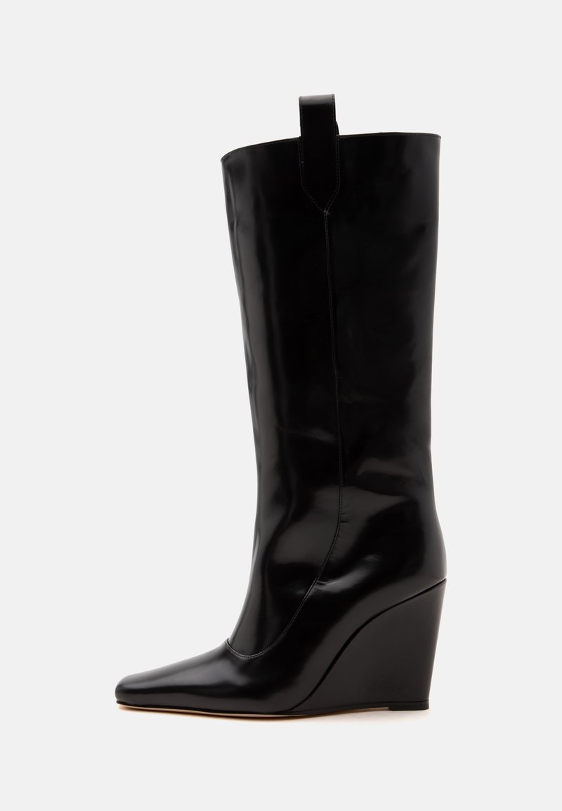 Botte compensée noire montante jusqu'au genou avec bout pointu et languette sur une tige en cuir lisse sur fond blanc.