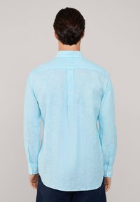 Camisa de manga larga a rayas azul claro, confeccionada en una tela suave, con un cuello abotonado y un dobladillo redondeado que presenta un detalle de pliegue en la parte posterior.