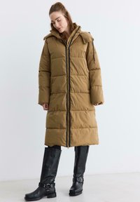 Brązowa kurtka puchowa o pikowanym wzorze, z zamkiem z przodu i kapturem. W połączeniu z czarnymi nadkolanówkami z klamrami.