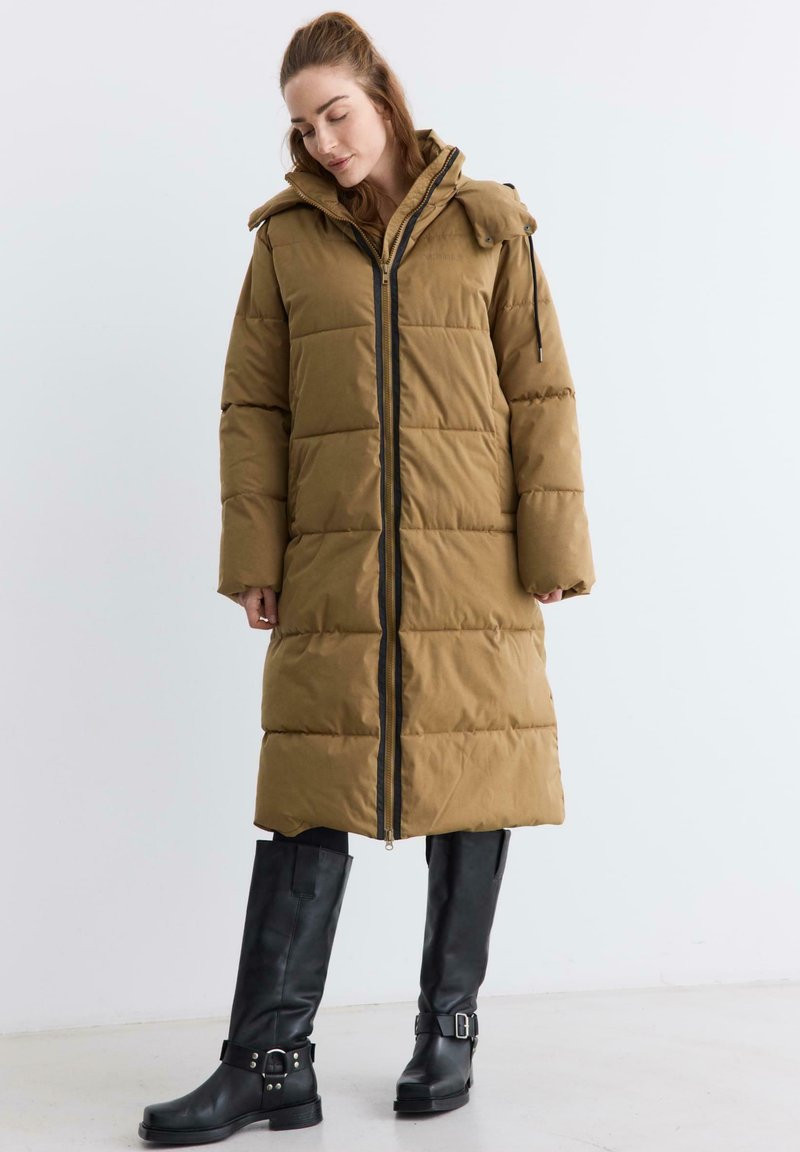 Brązowa kurtka puchowa o pikowanym wzorze, z zamkiem z przodu i kapturem. W połączeniu z czarnymi nadkolanówkami z klamrami.