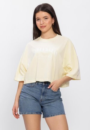 Jonge vrouw die een losse, oversized crème T-shirt draagt en een hoog getailleerde blauwe denimshorts, poserend met één hand in de zak.