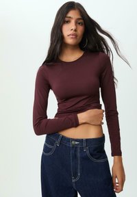 Maglietta a maniche lunghe color bordeaux con un design aderente e scollatura rotonda, abbinata a jeans in denim scuro a vita alta con dettagli di cuciture bianche.