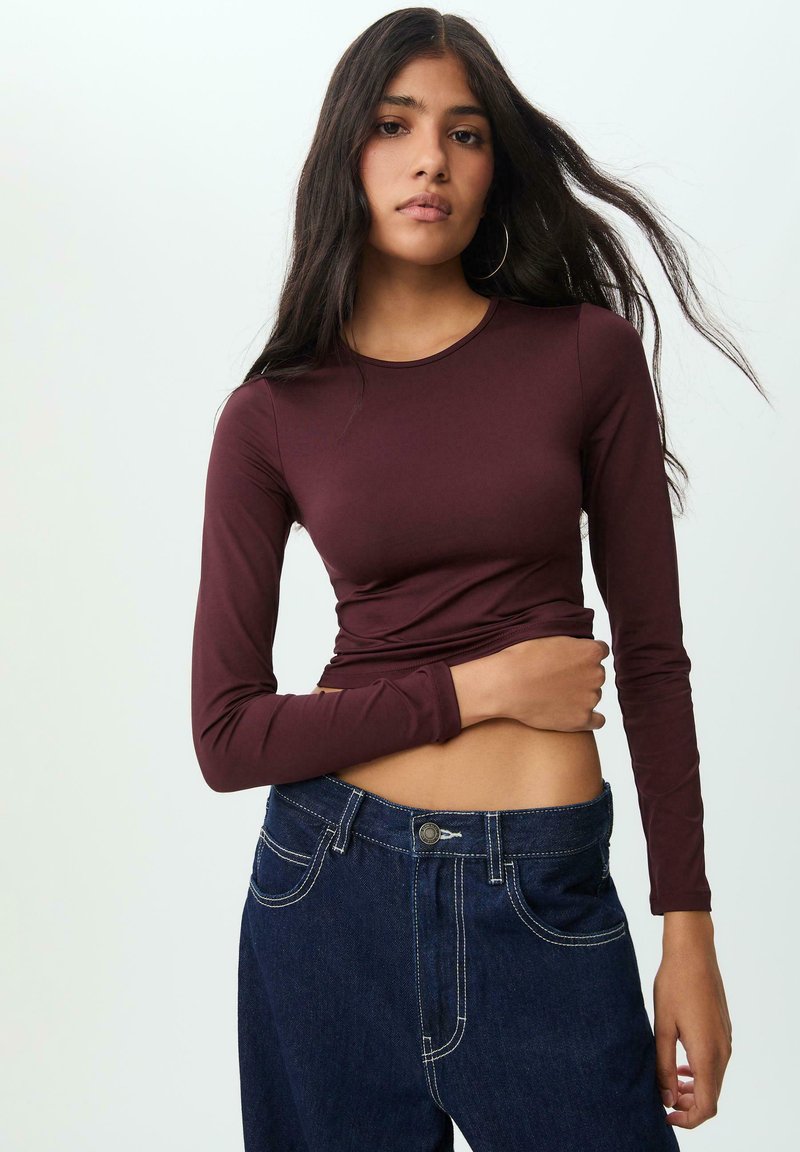 Maglietta a maniche lunghe color bordeaux con un design aderente e scollatura rotonda, abbinata a jeans in denim scuro a vita alta con dettagli di cuciture bianche.