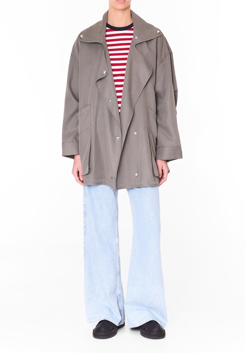 Une personne porte une veste grise surdimensionnée à grands revers, une chemise rayée rouge et blanche, un jean bleu clair à jambes larges et des chaussures noires.