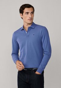 Camisa polo de manga larga en tejido azul claro, con cuello, tapeta de tres botones y logotipo bordado en el lado izquierdo del pecho.