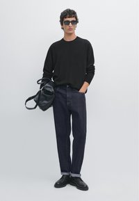 Czarny sweter, ciemne dżinsy z podwiniętymi mankietami, czarne sk�órzane buty i czarna skórzana torba. Kwadratowe okulary przeciwsłoneczne; neutralne tło.