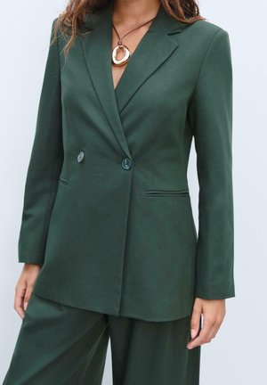 Femme portant un blazer vert foncé à double boutonnage et un pantalon assorti, avec un grand pendentif rond sur un cordon.