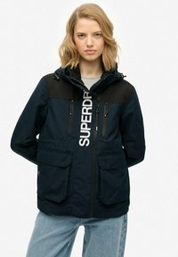 Granatowa kurtka z czarnymi akcentami, kapturem, zamkiem z przodu i dwiema dużymi kieszeniami, z białym logo "SUPERDRY" na przodzie. Gładka faktura.