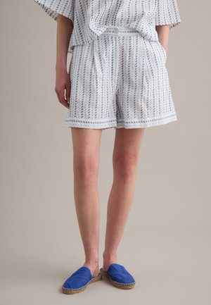 Persoon die wit-blauwe gestreepte shorts draagt met een bijpassende top en blauwe slip-on espadrilles, staand tegen een neutrale achtergrond.