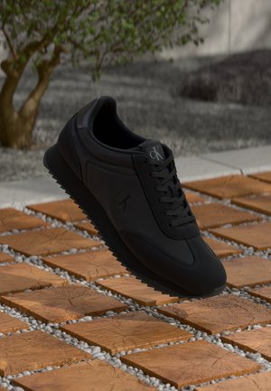 Zwarte Calvin Klein sneaker met veters zwevend boven houten vierkante tegels en grind, met een klein boompje op de achtergrond.