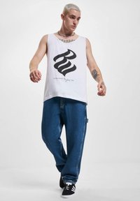 Un maiou alb cu un grafic mare negru și text, asociat cu pantaloni denim albaștri lejeri. Modelul poartă adidași negri cu dungi albe.