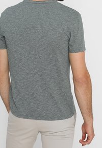 Mężczyzna w szarej koszulce z krótkim rękawem i jasnobeżowych spodniach, pokazany z tyłu na jednolitym tle.