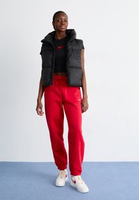 Gilet imbottito nero con collo alto, t-shirt rossa con logo Nike, pantaloni della tuta rossi e sneakers bianche con accenti rossi contro uno sfondo neutro.