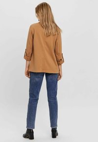 Hnědý top s dlouhými rukávy a ohrnutými manžetami, kombinovaný s modrými džínami. Top má volný střih a hladkou texturu látky. Černé boty na podpatku.