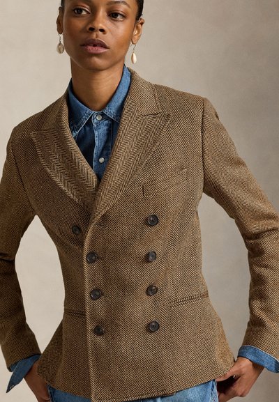 Καφέ σακάκι διπλής σειράς κουμπιών με σχέδιο ψαροκόκκαλο και μαύρα κουμπιά, σε συνδυασμό με πουκάμισο τζιν, που αναδεικνύει εφαρμοστή γραμμή και υφή υφάσματος.