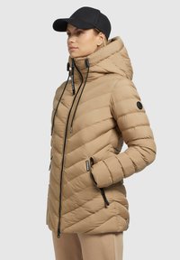 Doudoune beige avec motif en chevrons, col haut, poches zippées et capuche ajustable. Accents noirs sur les fermetures éclair et les manches.
