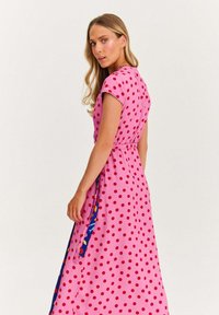 Žena s dlouhými blond vlasy, oblečená v růžových šatech s červenými puntíky, stojící na bílém pozadí a dívající se přes rameno.