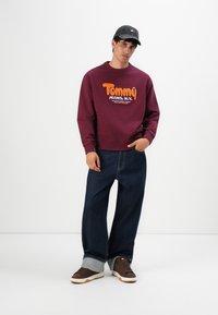 Joven con un sudadera burdeos de Tommy Jeans, pantalones oscuros de pierna ancha con grandes dobladillos, zapatillas marrones y una gorra de béisbol oscura, de pie.