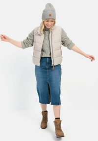 Helle beige Pufferweste, grauer Rippstrickpullover, blauer Jeansrock mit Schlitz vorne, braune Schnürstiefel, graue Mütze mit Logo.