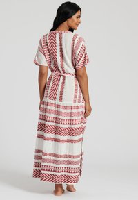 Maxiklänning i vitt tyg med djärva röda geometriska mönster. Har korta ärmar, en åtsittande midja och en flerlagrad volangkant.