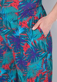 Tessuto stampato tropicale colorato in tonalità di rosso, teal e viola; presenta una tasca e un materiale leggero e testurizzato.