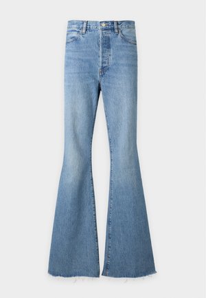 Femme portant un haut côtelé jaune pâle à manches longues avec des découpes diagonales et détails à boutons, associé à un jean bleu taille haute à jambes larges.