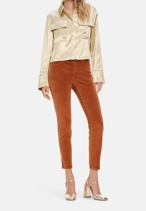 Mujer con camisa brillante beige de manga larga, pantalones ajustados color óxido y zapatos de tacón alto dorados con tiras en el tobillo, de pie sobre fondo blanco.