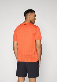 Camiseta deportiva de manga corta en naranja brillante con rayas verticales texturizadas y corte regular. Logo en la parte posterior del cuello. Combinada con pantalones cortos negros.