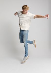 Lichtbeige kortarm overhemd, lichtblauwe spijkerbroek en grijze sneakers met witte strepen. Model in een speelse pose tegen een effen achtergrond.