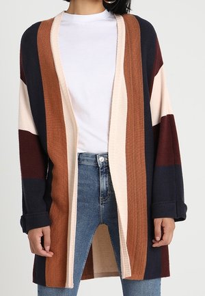 Frau trägt eine offene Strickjacke mit breiten vertikalen Streifen in Marineblau, Braun, Creme und Burgunderrot über einem weißen Hemd und blauen Jeans.