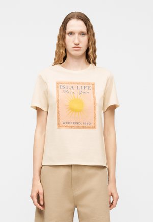 Hajlított hajú nő, aki bézs színű pólót visel, melyen sárga nap és az "ISLA LIFE Ibiza, Spanyolország HÉTVÉGE, 1983" felirat látható, valamint bézs rövidnadrágot.