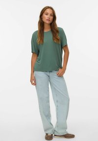 Oversized, groene top met korte mouwen en een ronde hals, gecombineerd met lichtblauwe wijde jeans en bruine veterschoenen.