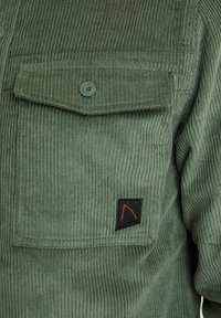 Vihreä corduroy-nappipaita rintataskulla, jossa on musta kangasmerkki oranssilla kuviolla ja kankaassa poikkijuovainen rakenne.