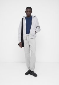 Sweat-shirt à capuche gris, t-shirt bleu, pantalon de survêtement gris et chaussures noires. Le modèle tient un sac tressé noir. Tissus doux et décontractés, coupe ample.