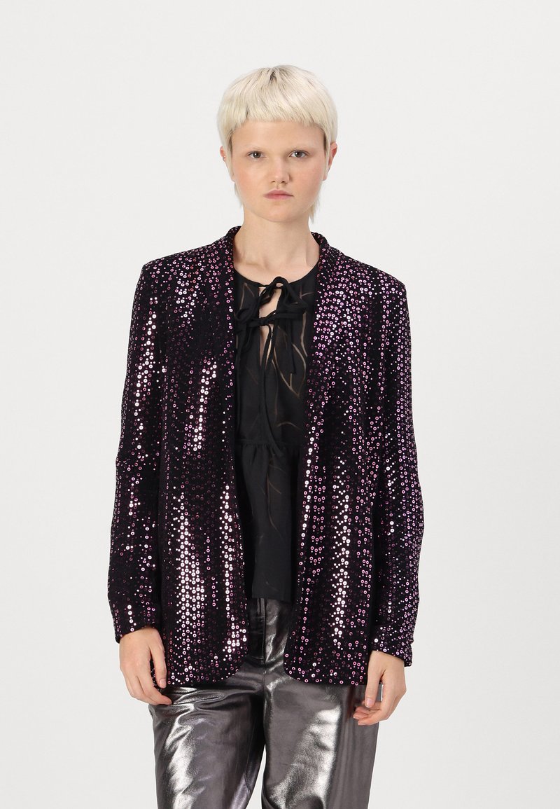 Veste en sequins de couleur violet foncé avec une texture brillante, devant ouvert et détail de nœud papillon noir. Associée à un haut noir et un pantalon argent métallique.