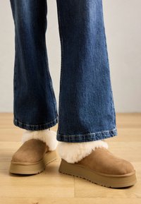 Pantofole in suede marrone con un spesso risvolto di pelliccia chiara, montate su una suola piatta beige, abbinate a jeans in denim a gamba larga.
