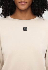 Felpa beige con scollo rotondo, texture morbida e una piccola etichetta nera sul petto. Design classico e minimalista.