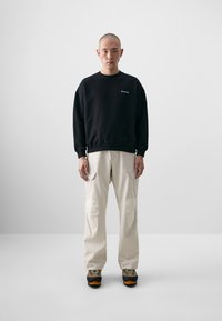 Sweatshirt en coton noir avec col rond et logo brodé, associé à un pantalon cargo beige avec des patchs aux genoux et des poches, et des chaussures grises.