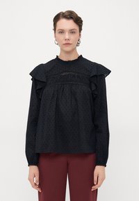 Blusa nera con pois a rilievo, maniche a volant e dettagli in pizzo. Presenta un collo alto e una vestibilità morbida, abbinata a pantaloni borgogna.