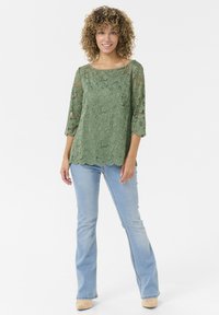 Top verde de encaje con patrón floral, mangas tres cuartos y dobladillo con forma de concha. Combinado con jeans acampanados de color azul claro y calzado beige.