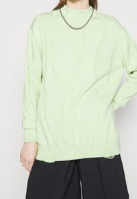 Ljusgrön tröja med ribbad, mock neck, texturerade vågmönster och fransiga fållar. Kombinerad med lösa svarta byxor.